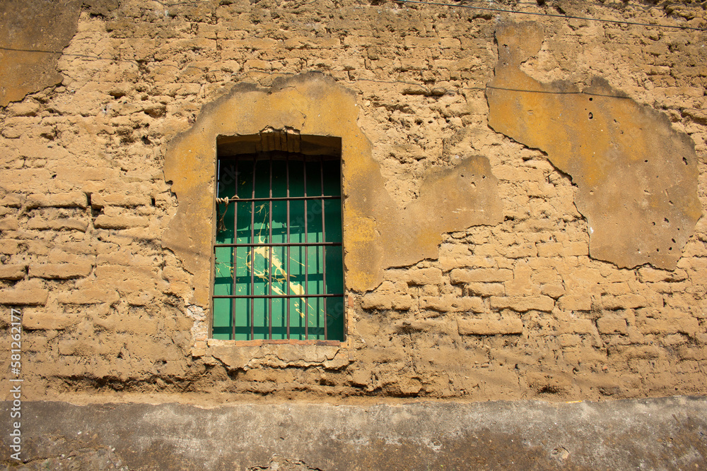 Ventana con barrotes en pared de adobe, pared rural de casa de campo en pueblo Ventana con barrotes en pared de adobe, pared rural de casa de campo en pueblo