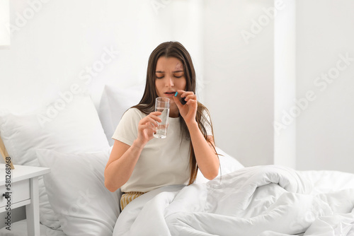 Young woman with glass of water taking vitamin supplement in bedroom