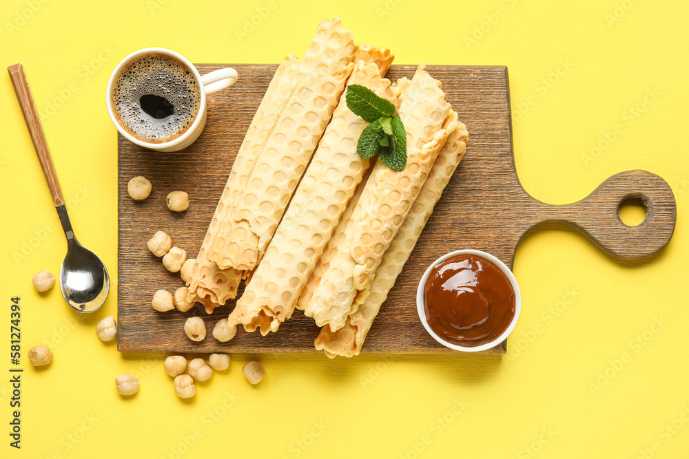 Board with delicious wafer rolls, boiled condensed milk, cup of coffee and hazelnuts on yellow background