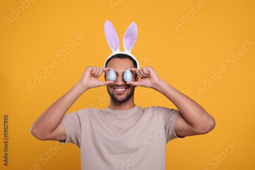 Happy African American man in bunny ears headband covering eyes with Easter eggs on orange background