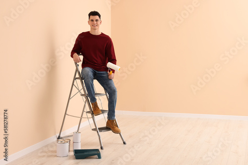 Young handsome man with roller on metal stepladder indoors, space for text. Room renovation
