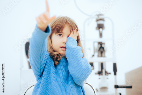 Cute Little Girl Covering One Eye During Ophthalmological Consult. Toddler child pointing to a vision chart during eye check
 photo