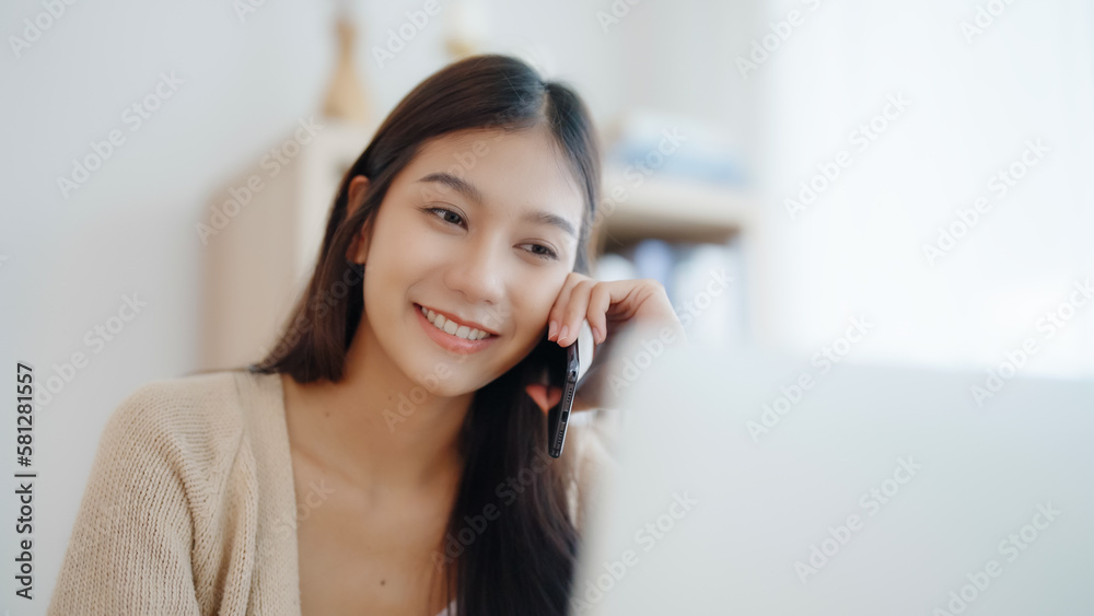 Young asian woman talking mobile smartphone while working on computer laptop on desk in living room at home Young asian woman talking mobile smartphone while working on computer laptop on desk in living room at home