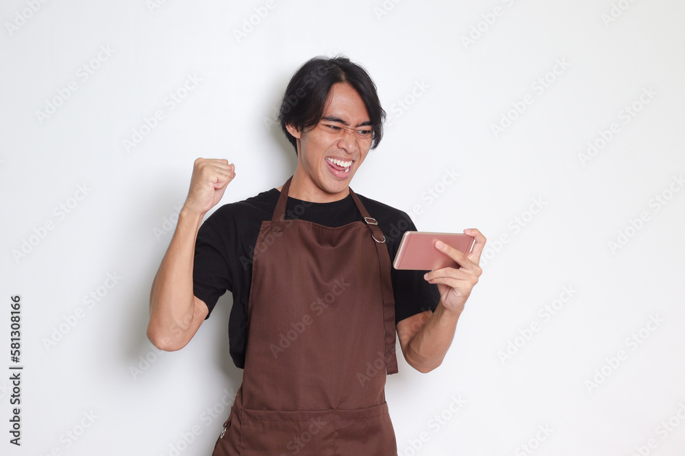 Portrait of attractive Asian barista man in brown apron holding mobile phone and watching video while raising his fist celebrating success. Isolated image on white background Portrait of attractive Asian barista man in brown apron holding mobile phone and watching video while raising his fist celebrating success. Isolated image on white background