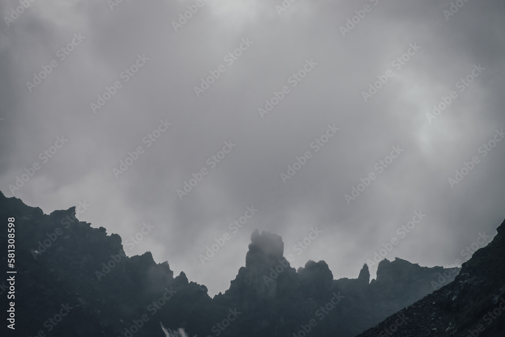 Dark atmospheric mountain landscape with black pointy rocky peak in gray cloudy sky. Lead gray low clouds among black mountains. Dark rocky pinnacle in low clouds in rainy weather. Gloomy minimalism. Dark atmospheric mountain landscape with black pointy rocky peak in gray cloudy sky. Lead gray low clouds among black mountains. Dark rocky pinnacle in low clouds in rainy weather. Gloomy minimalism.