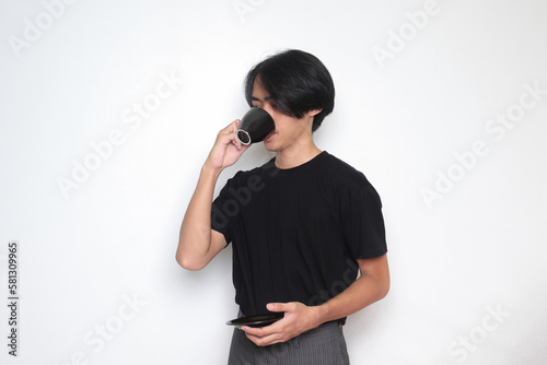Portrait of handsome Asian man in black t-shirt drinking a cup of coffee while holding the saucer. Isolated image on white background