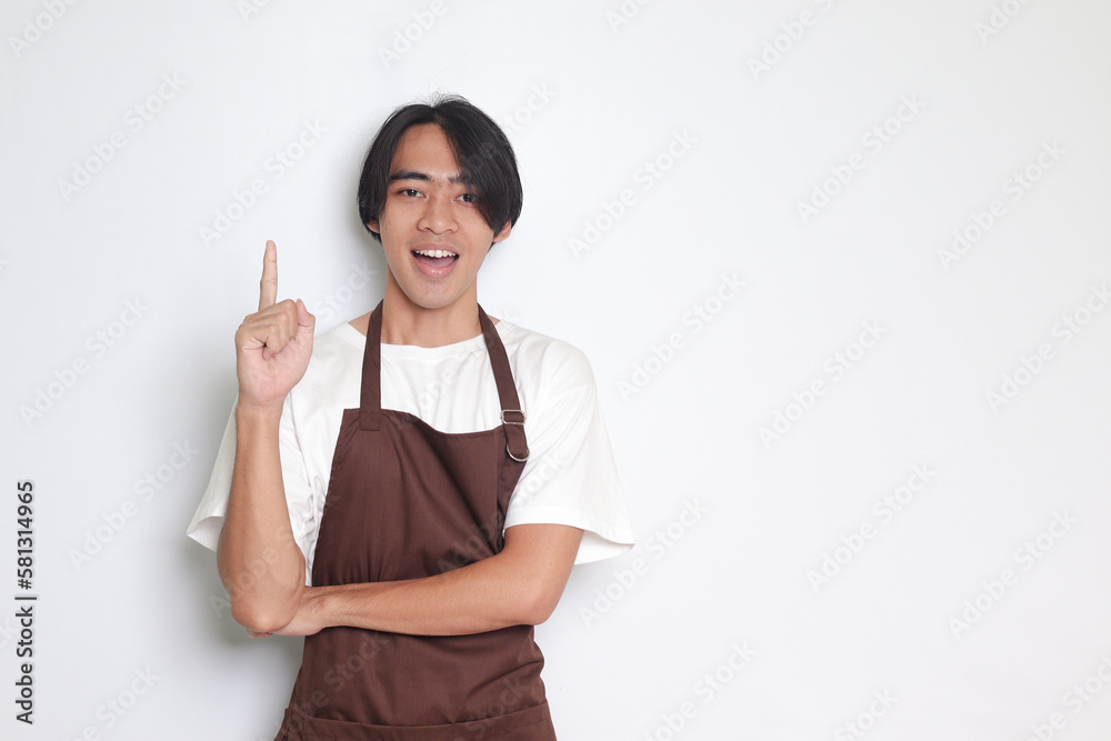 Portrait of attractive Asian barista man in brown apron showing product, pointing at something with hands. Advertising concept. Isolated image on white background