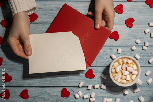 Female hands holding empty valentine card mock up red envelope with white cup of coffee and marshmallows on wooden blue background. Romantic Small hearts Valentine day. Blank paper card copy space for