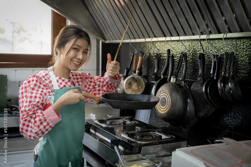 Φωτογραφία Beautiful asian home cook girl wearing apron and holding wooden spatula and frying pan while standing in kitchen background with smile on her face