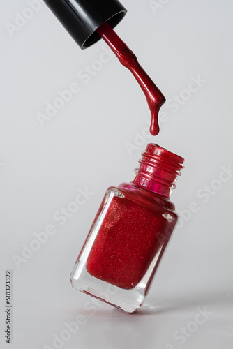 A bottle of bright red nail polish stands on a light table. A drop of varnish hangs from the brush. Light background