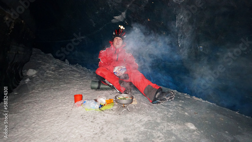 A guy cooks food on gas inside an ice cave. The climber lights the burner, cuts sausage and tomato. Breaks the eggs and fry in a frying pan. Steam from food. The ice wall is blue and the floor is gray photo