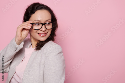 Confident portrait of a beautiful positive multi-ethnic woman, wearing stylish black frame glasses, smiling looking aside at copy advertising space over isolated pik background. People. Optics. Style