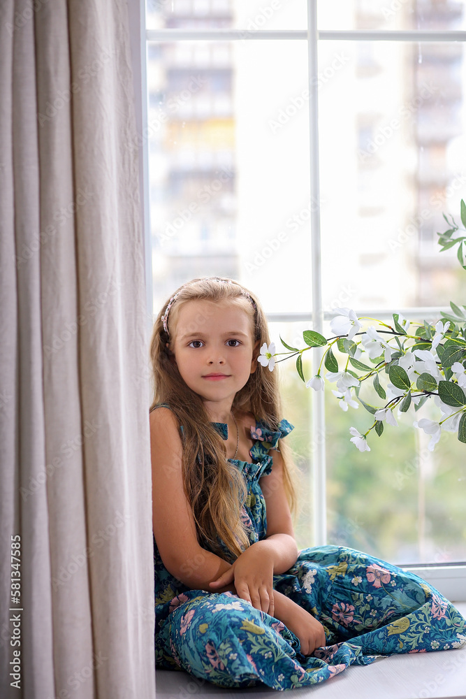Cute baby girl Caucasian in a green floral sundress sitting on the windowsill by the window. City in the background. Home comfort concept. Cute baby girl Caucasian in a green floral sundress sitting on the windowsill by the window. City in the background. Home comfort concept.