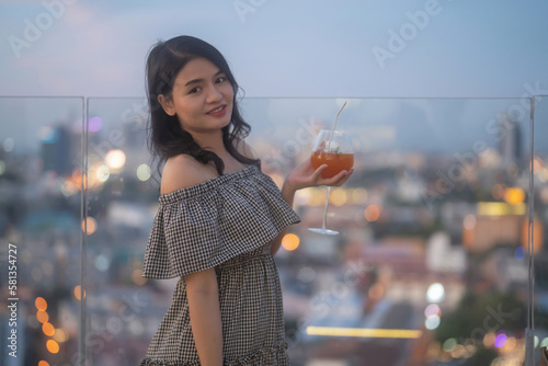 Portrait of an Asian woman holding a cocktail beer alcohol drink glass at night in restaurant on balcony sky bar with urban city skyline background. Sweet fruit food. People lifestyle.