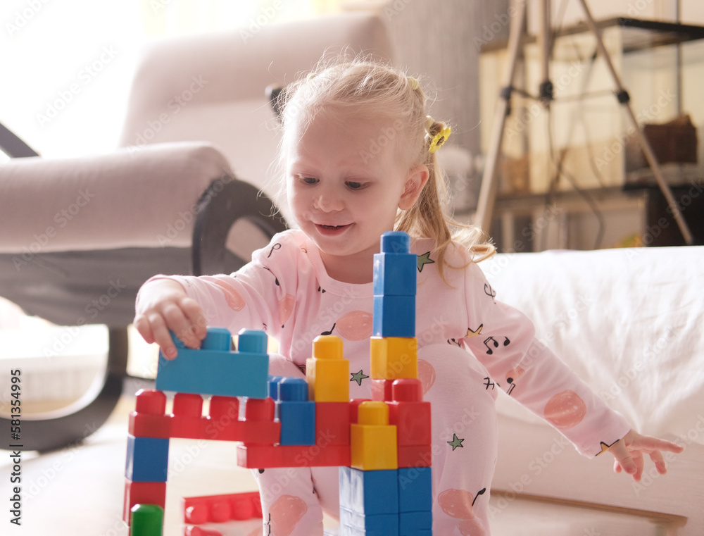 Naklejka premium A laughing little child, a blonde preschooler playing with colorful cubes, sitting on the floor in pink pajamas, in a sunny room with a large window at home. Early development of children.