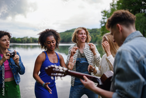 A group of young friends having fun near a lake, laughing and playing guitar.