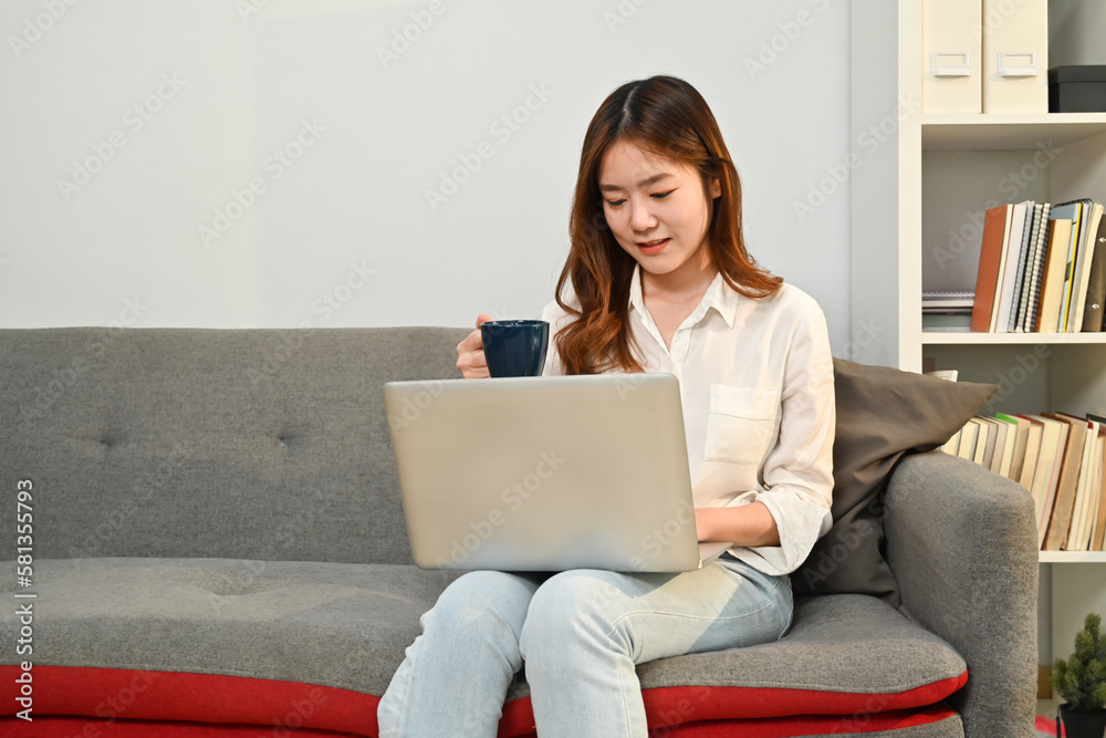 Pretty young asian woman drinking coffee and working remotely from home, using laptop on couch Pretty young asian woman drinking coffee and working remotely from home, using laptop on couch