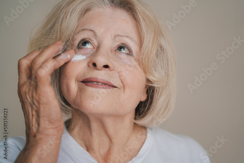 Smiling old caucasian woman applying anti aging cream on her face against grey background. Beauty portrait. Facial treatment. Anti-wrinkles care. Anti-aging