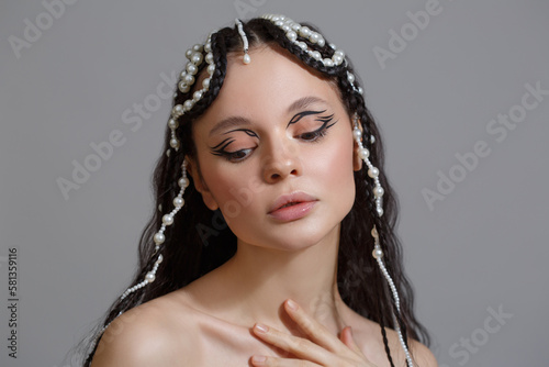 Beauty portrait of a brunette girl with a braid hairstyle with pearls on long hair isolated on a gray background.