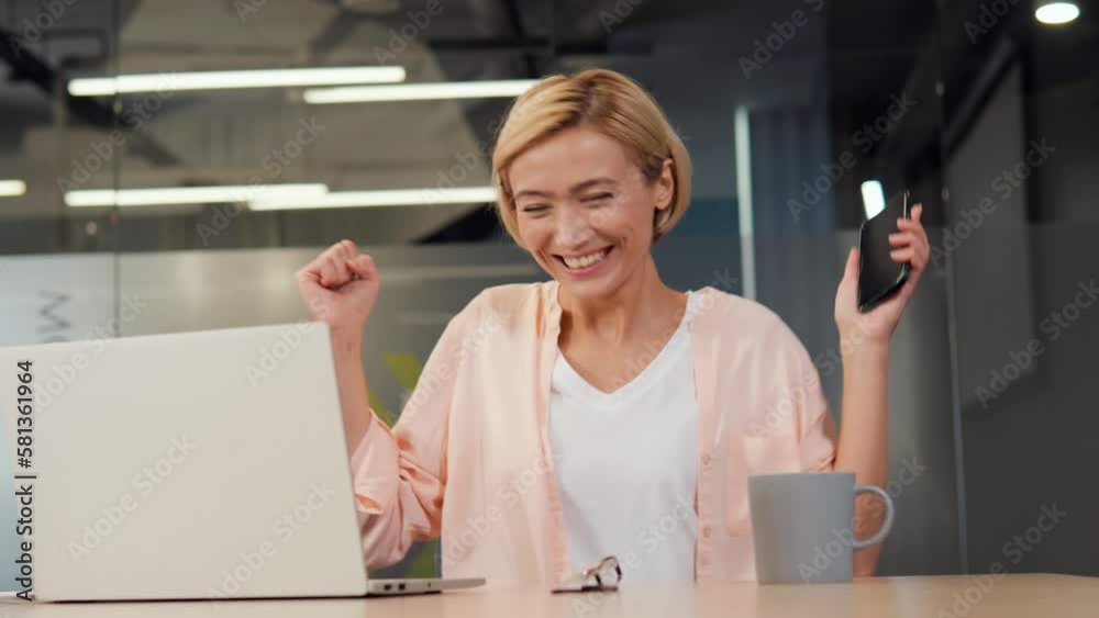 Amazing woman is seated at desk, holding cellphone received SMS from bank and is feeling incredibly happy. Woman makes yes I did it gesture looks very excited. Achievement, winning a lottery prize. Amazing woman is seated at desk, holding cellphone received SMS from bank and is feeling incredibly happy. Woman makes yes I did it gesture looks very excited. Achievement, winning a lottery prize.