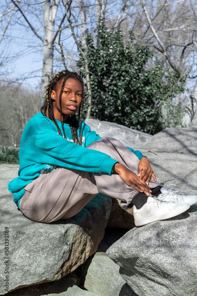 Obraz premium Mujer joven de color con trenzas en el pelo vestida con un chándal sentada en las piedras con la mirada perdida de un parque de Madrid 