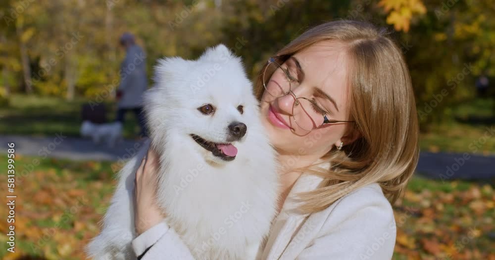 Blonde-haired woman in glasses strokes fluffy dog head holding in arms in autumn park. White pet enjoys female owner care and walking in park illuminated by sun Blonde-haired woman in glasses strokes fluffy dog head holding in arms in autumn park. White pet enjoys female owner care and walking in park illuminated by sun