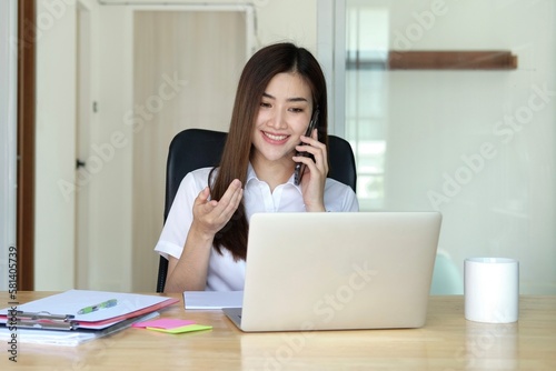 Smiling professional young Asian businesswoman talking in front of the camera having video conference with her business partners on laptop in a contemporary office space