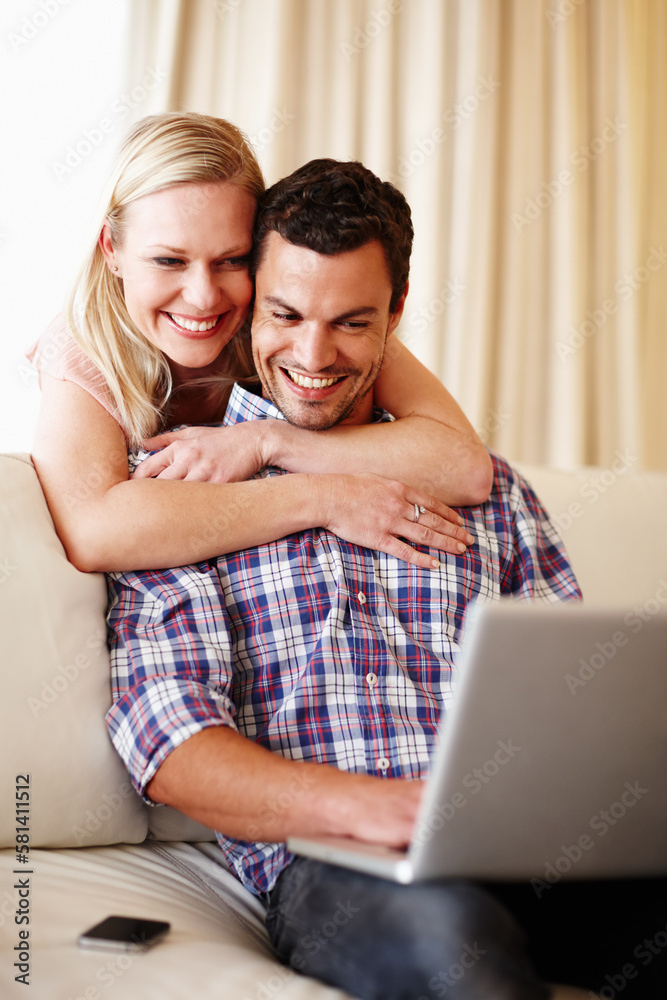 Sharing a funny video clip. A beautiful woman hugging her husband from behind while he sits on the couch working on his laptop.