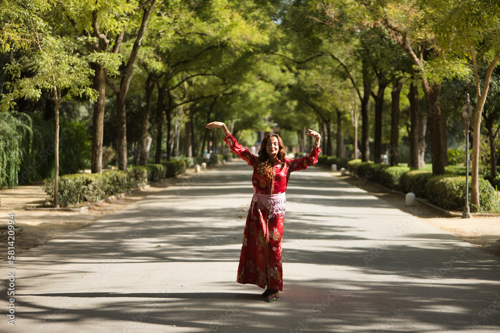 Beautiful young woman in a typical Moroccan red suit, embroidered with gold and silver threads, dancing in an outdoor park avenue. Concept beauty, ethnicity, typical suits, Marrakech, Arab.