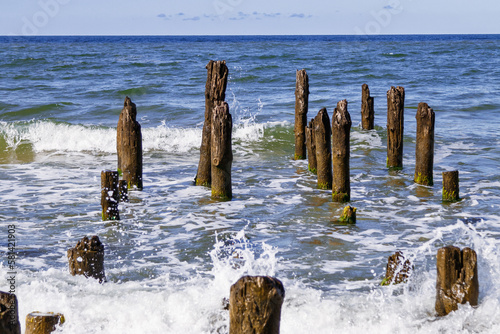 Wooden piles from old breakwater on sandy beach, blue sky, blue sea water of Baltic sea, Filinskaya bay beach, Russia. Weathered wooden poles in water. Summer day, waves and wind, landscape