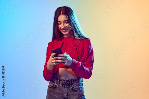 Pretty teen girl looking at cell phone over gradient blue-yellow background. Youth and virtual lifestyle of future. Concept of games, remoute work, technology. photo