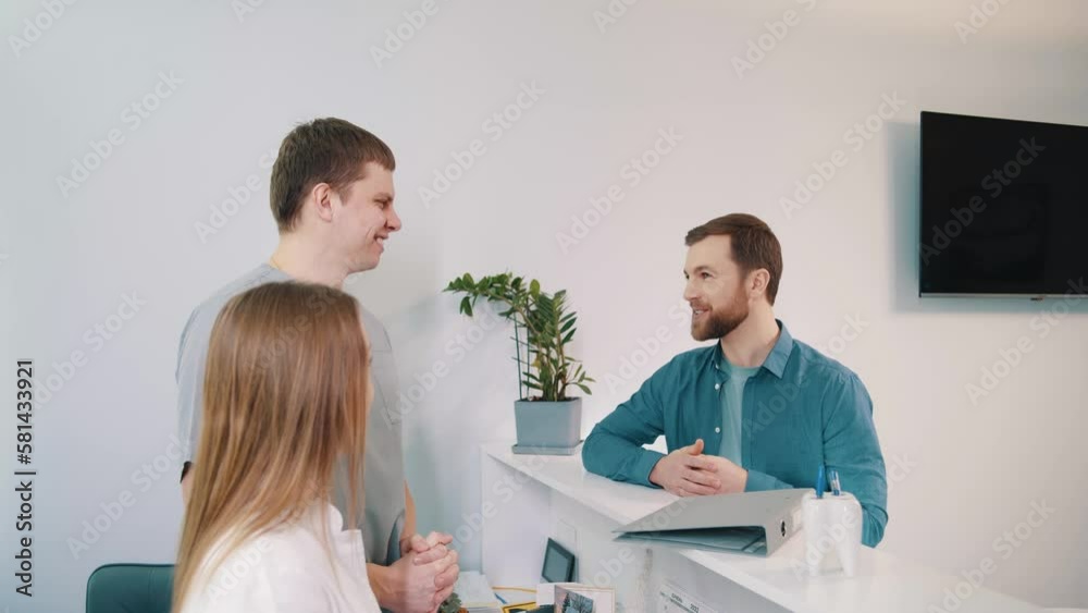 In a large modern clinic, a young smilling patient communicates with doctors on the reception desk and go to the check appointment to the doctor In a large modern clinic, a young smilling patient communicates with doctors on the reception desk and go to the check appointment to the doctor