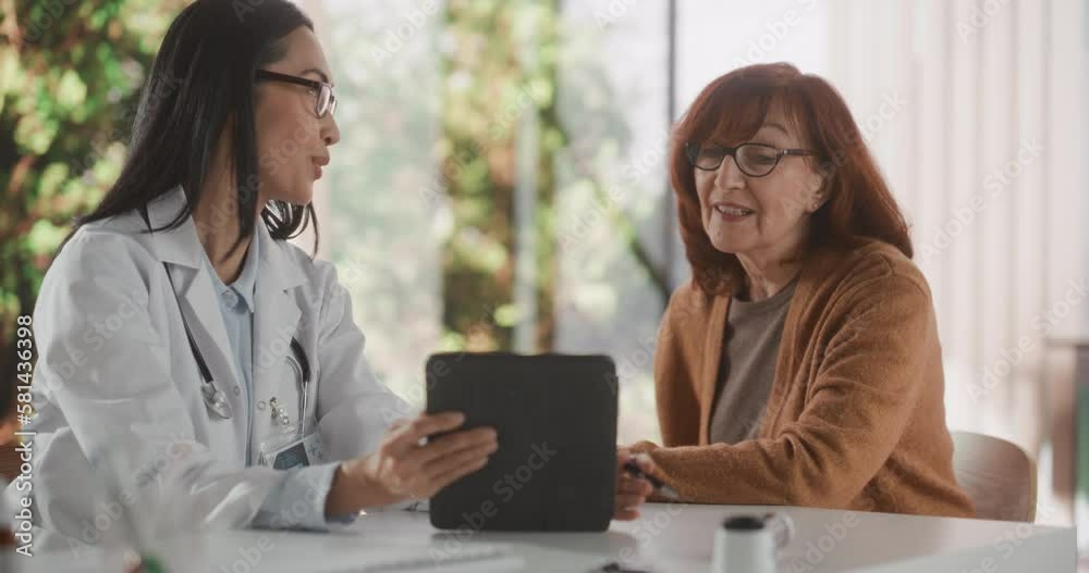 Female Physician Talking with a Senior Woman During Consultation in Health Clinic and Showing Her X-Ray Scans on Tablet Computer. Asian Nurse in White Coat Suggesting Treatment to Elderly Patient Female Physician Talking with a Senior Woman During Consultation in Health Clinic and Showing Her X-Ray Scans on Tablet Computer. Asian Nurse in White Coat Suggesting Treatment to Elderly Patient