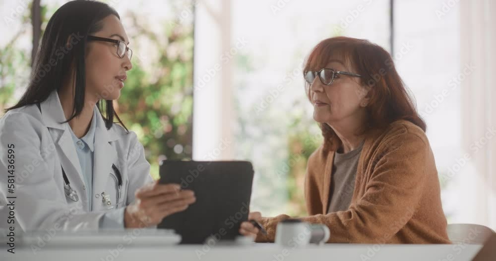 Young Asian Doctor Showing Heart Analysis Results on Tablet Computer to Senior Female Patient During Consultation in a Health Clinic. Physician Sitting Behind a Desk in Hospital Office