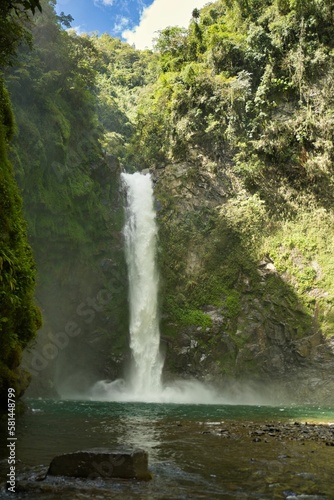 An imposing waterfall in an enclosed valley in Banaue in the Philippines that flows down a large green hill and flows into a pool.