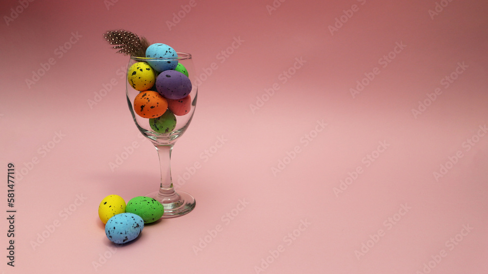 Colorful Easter eggs in a glass cup with a feather, on a pink background.