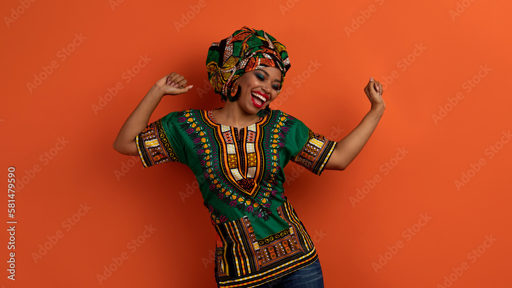Fototapeta premium Joyful pretty black woman in african costume dancing on orange