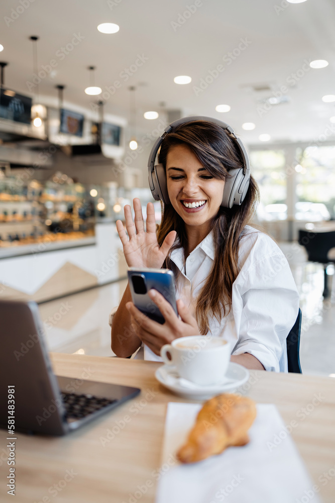 Beautiful and happy young woman sitting and eating delicious rolls in bakery or fast food. She also using her laptop and listening to music with headphones.