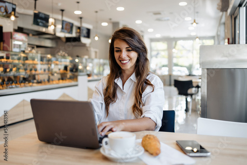Beautiful and happy young woman sitting and eating delicious rolls in bakery or fast food while she using laptop computer.