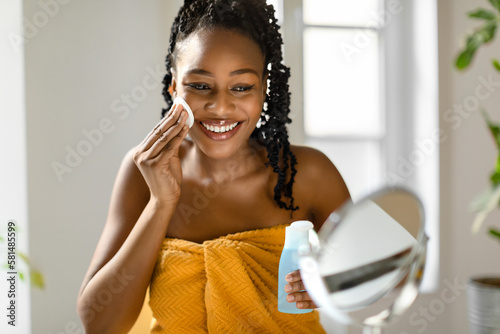 Face care routine. Young african american lady wrapped in towel holding bottle with micellar water and using cotton pad photo