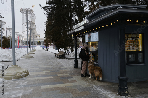 Young pretty woman buys coffee at fast food store in park in winter, dog standing next to and asks for yummy too. Girl on walk with Australian Shepherd. Dog friendly cafe shop. Full-length portrait.
