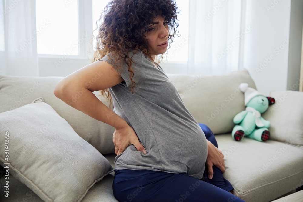 Pregnant woman sitting on the couch lower back pain and headache, strain on the spine during pregnancy. Lifestyle preparation for childbirth, last month of pregnancy Pregnant woman sitting on the couch lower back pain and headache, strain on the spine during pregnancy. Lifestyle preparation for childbirth, last month of pregnancy