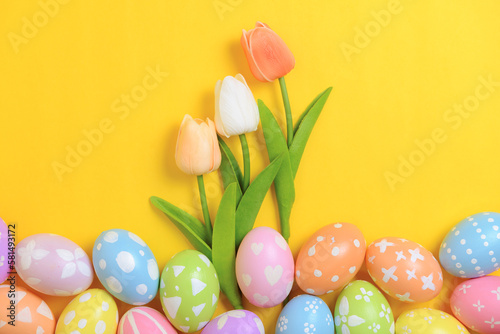 Happy Easter holiday greeting card design concept. Colorful Easter Eggs and spring flowers on yellow background. Flat lay, top view, copy space.