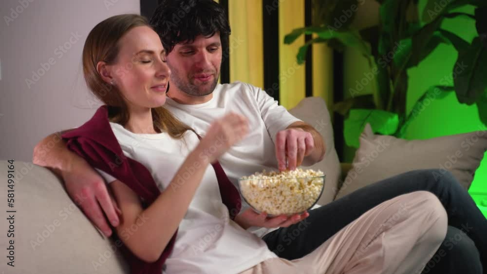 Charming couple who eats popcorn from a cup while sitting on the couch indoors and watching movies. Charming couple who eats popcorn from a cup while sitting on the couch indoors and watching movies.