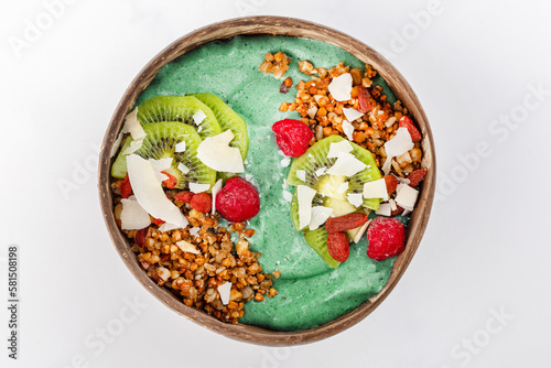 Green smoothie bowl in a bowl of coconut with yogurt, raspberries, coconut, kiwi, raspberries, nuts and roasted buckwheat. Top view, white background.