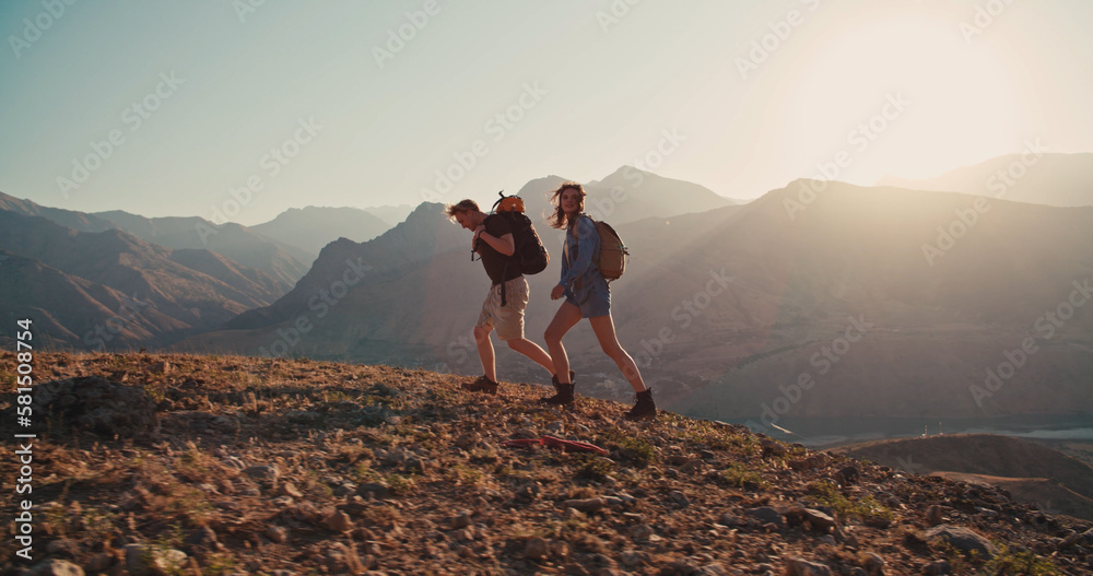 Couple of Young Happy Travelers Hiking with Backpacks on the Beautiful Rocky Trail at Warm Sunny Evening. Family Travel and Adventure Concept.