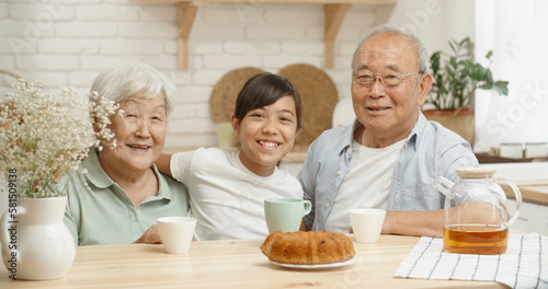 Mature asian couple and their teen granddaughter sitting together at kitchen table  smiling and looking at camera - family ties concept portrait closeup 