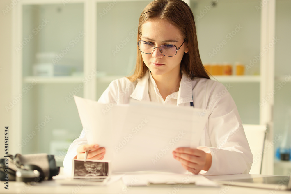 Young doctor in glasses reads medical documents while sitting at table in medical clinic. Young doctor in glasses reads medical documents while sitting at table in medical clinic.
