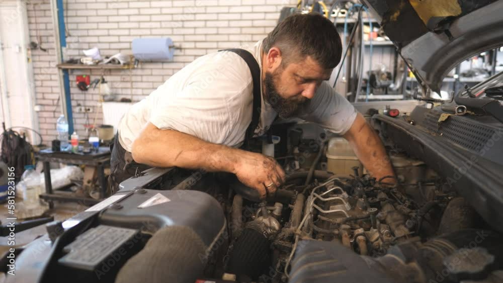 Repairman fixing engine of a car at workshop or service. Professional auto mechanic works under hood of vehicle. Bearded man checking automobile motor at garage. Concept of maintenance and diagnostic Repairman fixing engine of a car at workshop or service. Professional auto mechanic works under hood of vehicle. Bearded man checking automobile motor at garage. Concept of maintenance and diagnostic