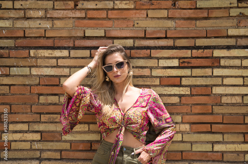 Young, blonde, beautiful woman with sunglasses is posing on red brick background while touching her hair and making different body expressions. The woman is on holiday. Beauty and fashion concept.